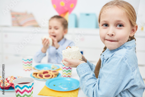Cute girl with large piece of birthday cake looking at camera while sitting by festive table