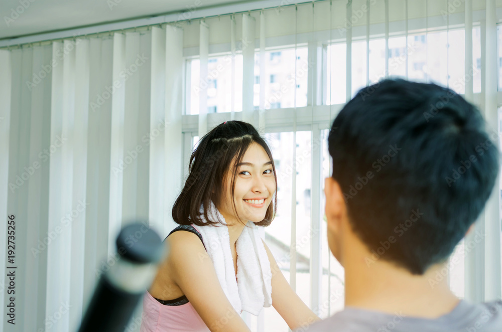 selective focus young happy smile asian woman slim body looking into camera and exercising in fitness with back view of personal trainer man gym for good healthy at the morning, sport concept selective focus young happy smile asian woman slim body looking into camera and exercising in fitness with back view of personal trainer man gym for good healthy at the morning, sport concept