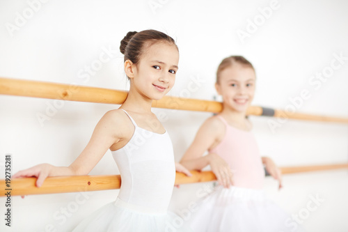 Happy little girl and her friend standing by wooden bars while training in ballet class