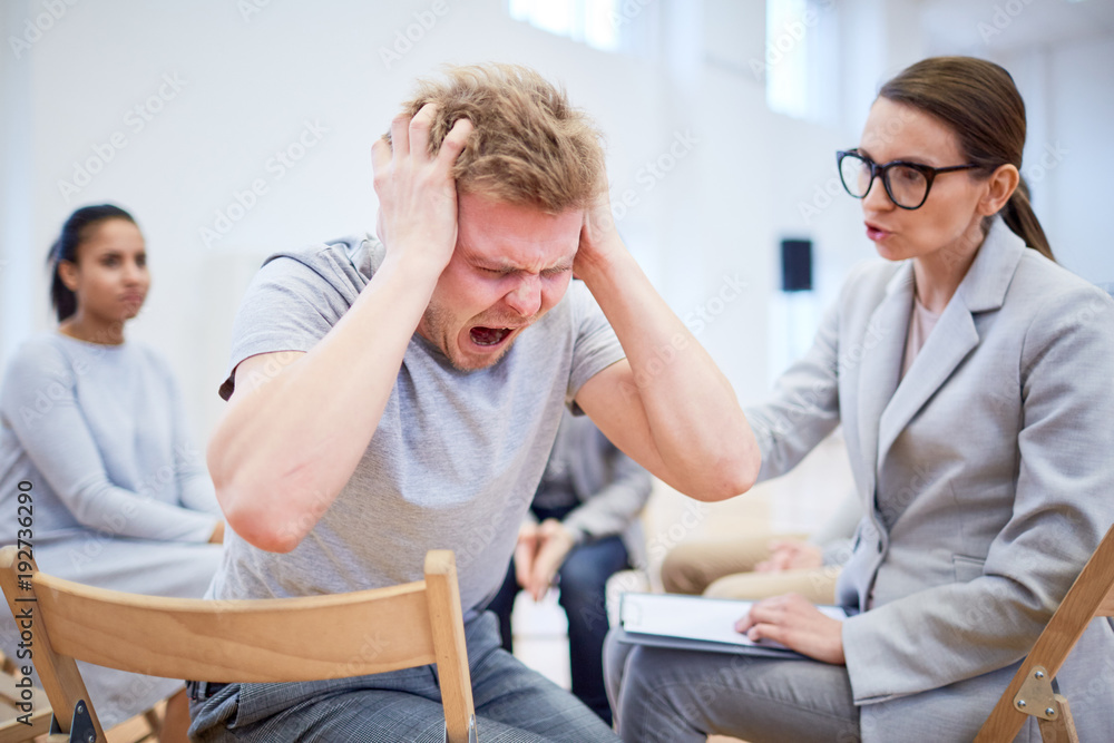 Troubled or stressed young man screaming while psychologist trying to support him during session Troubled or stressed young man screaming while psychologist trying to support him during session