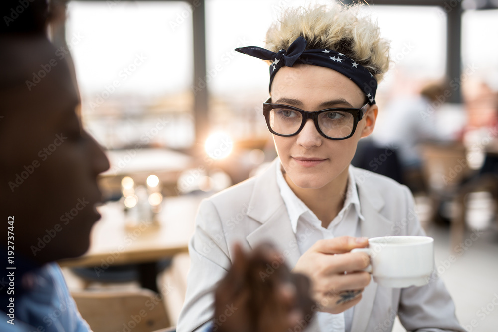 Serious woman in eyeglasses and headband having tea or coffee and listening to her colleague during conversation Serious woman in eyeglasses and headband having tea or coffee and listening to her colleague during conversation