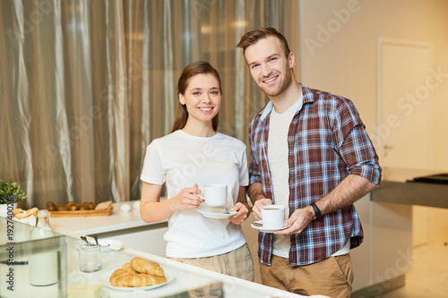 Young man and woman with cups of tea looking at camera in the kitchen at home