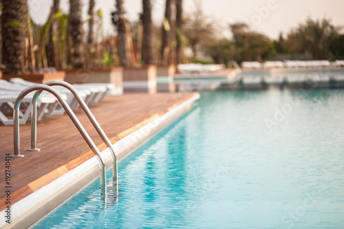 Empty hotel water pool with turquoise colour water at sunrise, focus on metal hand railing of staircase, copy space, place for text/ Relaxation, summer, vacation and water - concept/ 