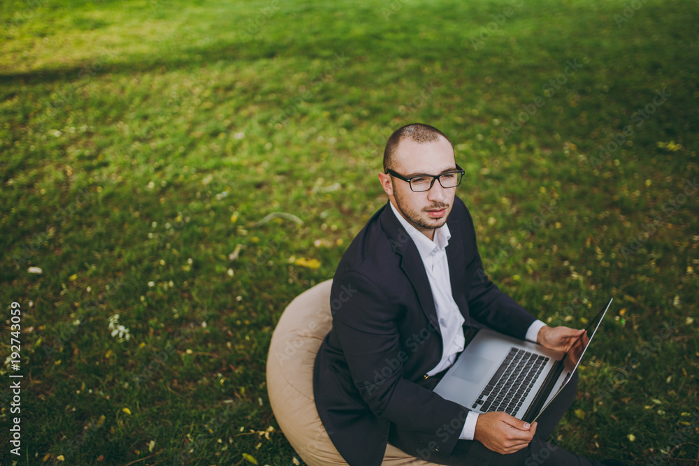Young successful businessman in white shirt, classic suit, glasses. Man sit on soft pouf, working on laptop pc computer in city park on green lawn outdoors on nature. Mobile Office concept. Top view. Young successful businessman in white shirt, classic suit, glasses. Man sit on soft pouf, working on laptop pc computer in city park on green lawn outdoors on nature. Mobile Office concept. Top view.