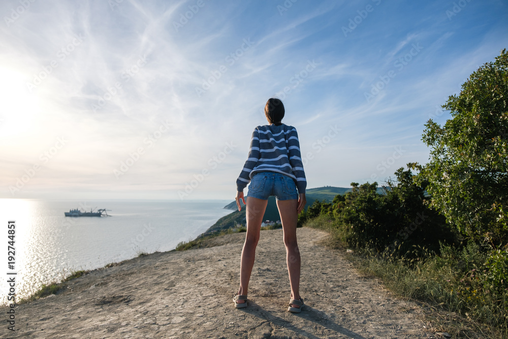 Unrecognizable woman in striped sweater and denim shorts stretching on the hill with seascape.