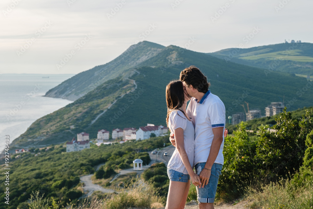 Naklejka premium Man kisses a woman on the cheek and hugs, standing on a mountain with seascape. Side view.