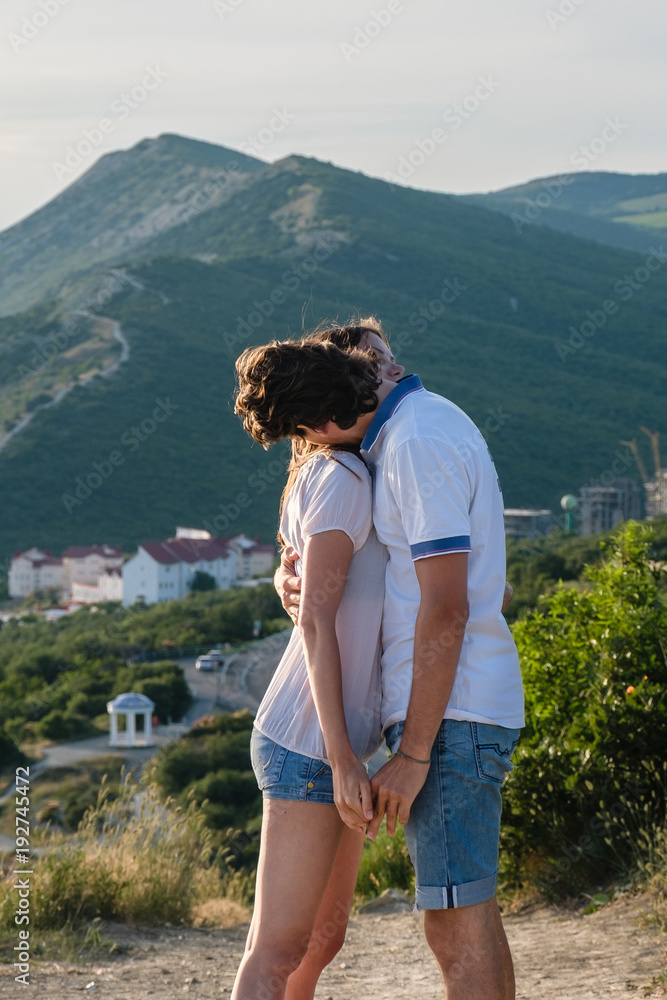 Man and woman in summer clothes cuddling standing on mountain with seascape at sunset. Side view.