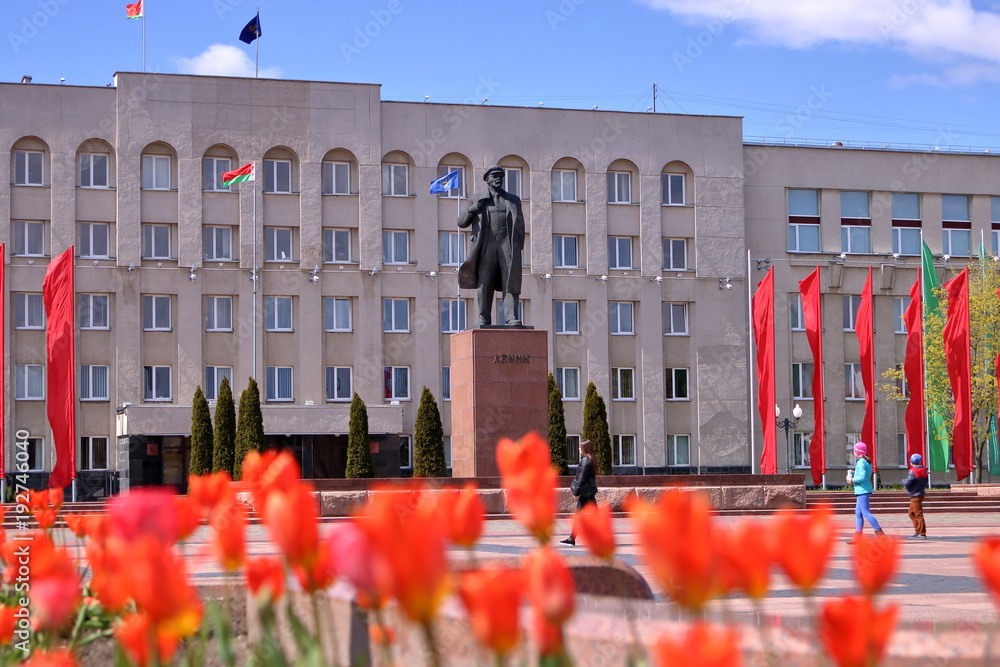 Pomnik Lenina na Białorusi, na pierwszym planie niewyraźne pomarańczowe tulipany, w tle budynek z wieloma oknami i flagą Białorusi, kilka osób na placu przed pomnikiem, na cokole cyrylicą LENIN - obrazy, fototapety, plakaty 