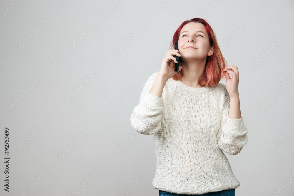 Portrait of pretty redhead girl wearing white sweater having phone conversation, smiling with teeth, posing against gray background with copy space. People, modern communication and technology Portrait of pretty redhead girl wearing white sweater having phone conversation, smiling with teeth, posing against gray background with copy space. People, modern communication and technology