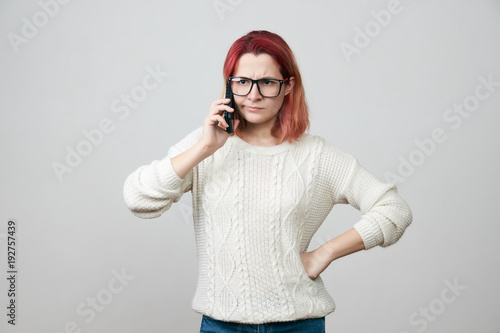Redhead woman model in glasses and white sweater complaining while talking on smartphone, confused and puzzled expression, frowning, being displeased. Girl retells situation to her friend. 