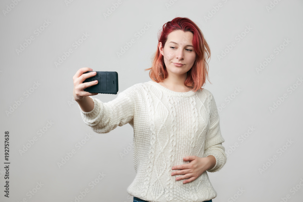 Naklejka premium Smiling young redhead woman with red hair making selfie using smart phone. Girl in white sweater taking photograph with mobile phone, looking with pretty smile at the camera of electronic device