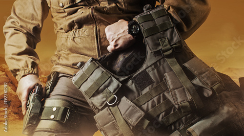 Photo of a soldier in military outfit holding a gun and bulletproof vest on orange desert background. photo