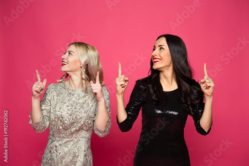 Friends forever. Close up photo of Two happy young beautiful smiling girlfriends in little black dresses posing and having fun on a pink background.