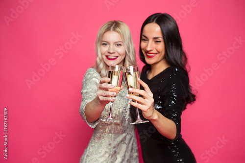 Two young beautiful smiling elegant women in evening dress and with glasses of champagne in their hands on a pink background.