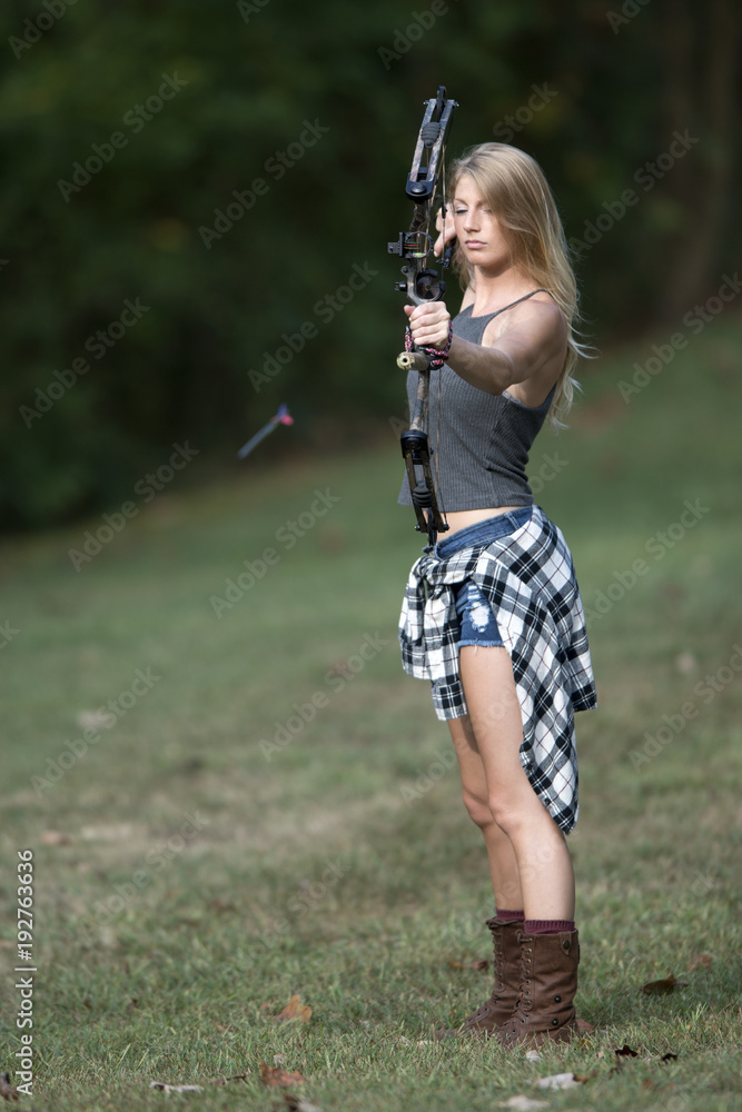 Stunning blonde female Caucasian archer shoots an arrow from a compound bow - arrow seen flying down range