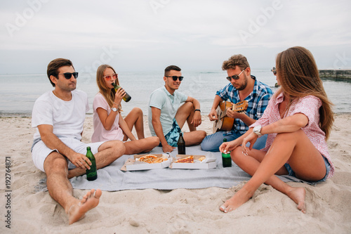 Group of young friends resting together on the beach with pizza and drink, sitting near the sea, listening song while one of them is playing guitar.