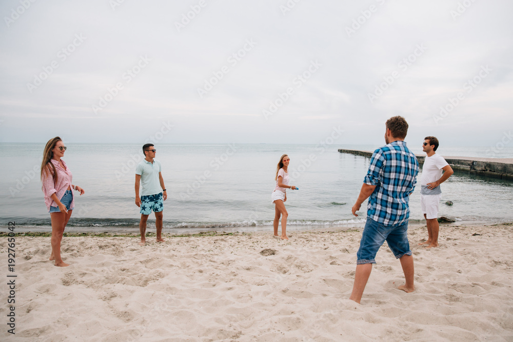 Group of young attractive friends playing frisbee on the beach, by the sea, spending weekend with pleasure.