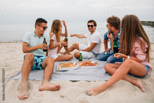 Photography Young company of attractive guys and girls relaxing together on the beach, sitting by the sea while drinking beer and eating pizza