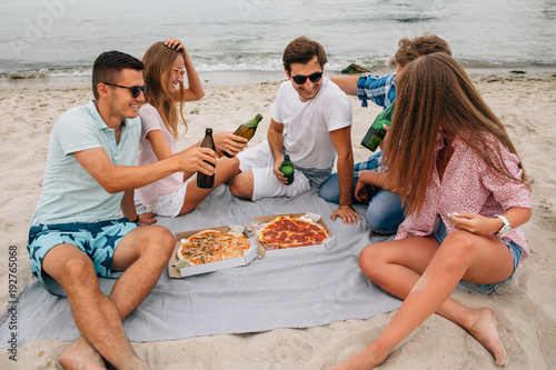 Young group of attractive friends resting on the beach, joking with each other, sitting by the sea, while drinking beer and eating pizza. © Maksym Azovtsev
