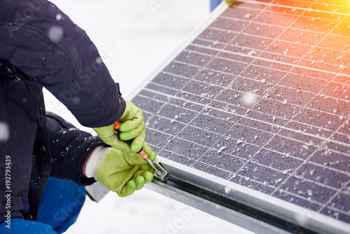 Hands of worker installing solar panels in snowy weather. Worker with tools maintaining photovoltaic panel. A bit of snow on solar panels. Close-up photo