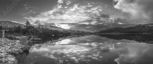 Stunning black and white sunrise landscape image in Winter of Llyn Cwellyn in Snowdonia National Park with snow capped mountains in background