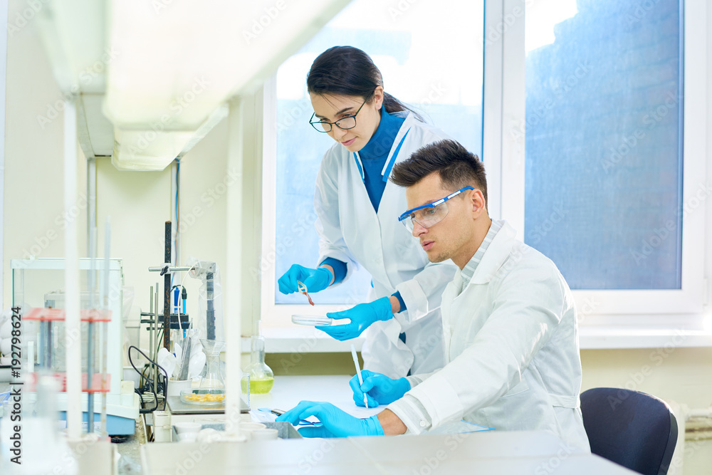 Team of ambitious researchers wearing white coats and rubber gloves carrying out experiment while gathered together at modern laboratory Team of ambitious researchers wearing white coats and rubber gloves carrying out experiment while gathered together at modern laboratory