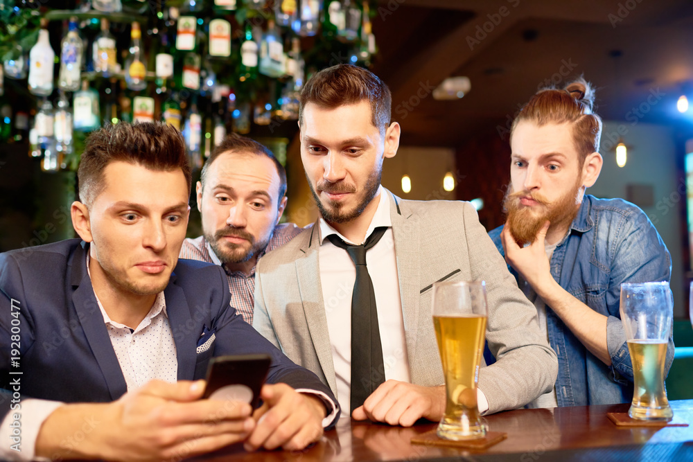 Group of surprised bearded men gathered together in bar, drinking lager and reading news with help of smartphone