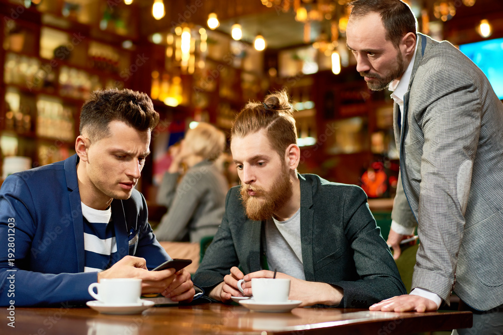 Handsome young manager using smartphone while having informal working meeting with colleagues, interior of modern cafe on background Handsome young manager using smartphone while having informal working meeting with colleagues, interior of modern cafe on background