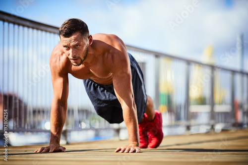 Determinated and athletic man doing pushups shirtless during workout session in the city