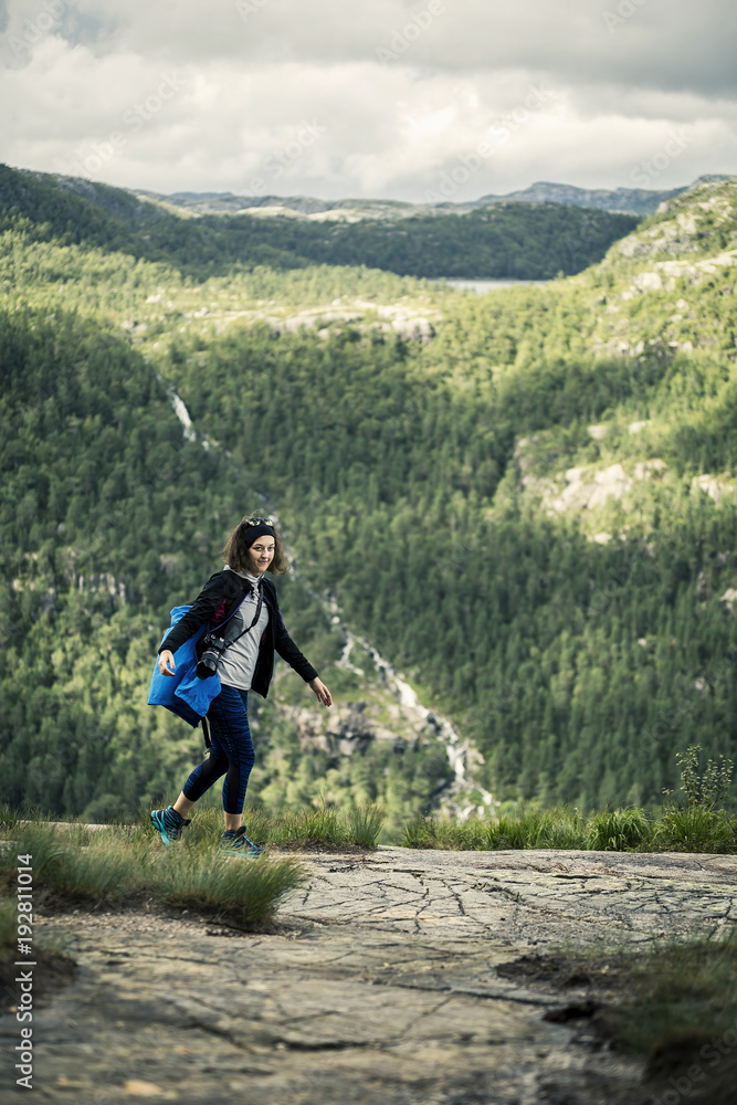 Portrait of young woman walking along the plateau in Preikestolen National park, Norway. She is looking at the camera, there is a green forest on background.