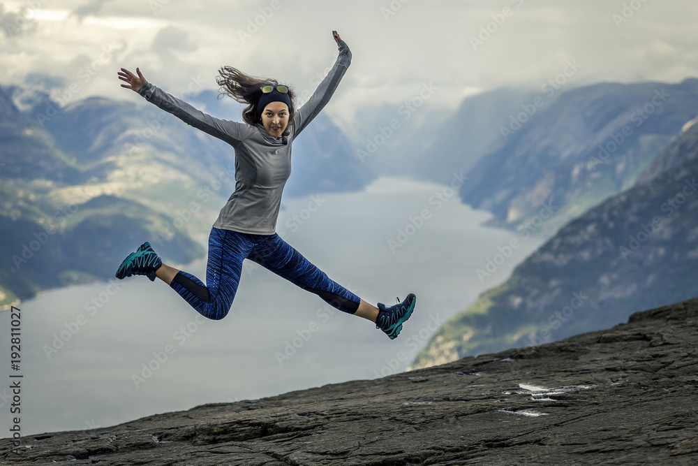 Portrait of young and sporty woman jumping on the Preikestolen plateau. Scenic fjord on background, Norway.