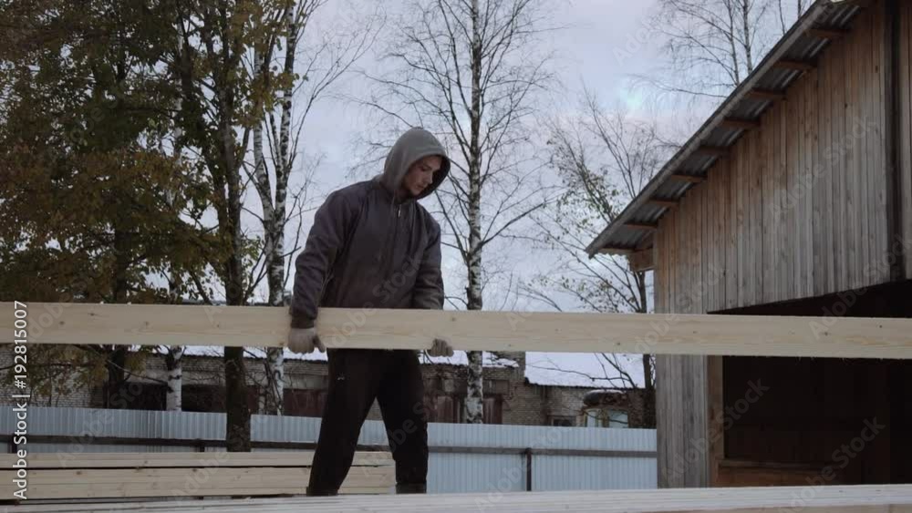 Caucasian man loader in dirty clothes carrying wood plank at sawmill factory yard on cold winter day Caucasian man loader in dirty clothes carrying wood plank at sawmill factory yard on cold winter day