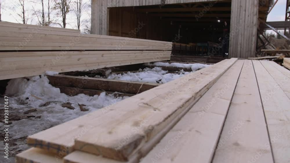 Man working at stacks of processed packed wooden planks stored in yard of sawmill, dirty ground with snow on sunny winter day Man working at stacks of processed packed wooden planks stored in yard of sawmill, dirty ground with snow on sunny winter day