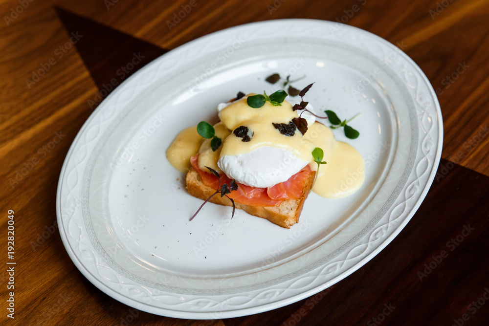 A horizontal image of eggs benedict on a white plate. A grilled toast with salmon, poached egg, hollandaise sauce, truffles and greenery. Selective focus. A horizontal image of eggs benedict on a white plate. A grilled toast with salmon, poached egg, hollandaise sauce, truffles and greenery. Selective focus.