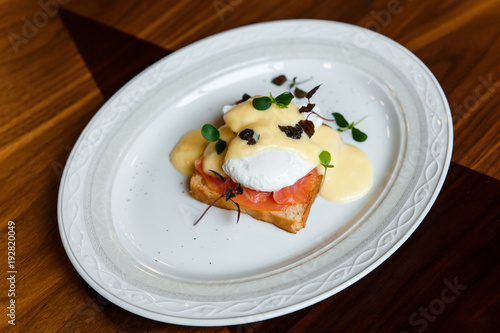 A horizontal image of eggs benedict on a white plate. A grilled toast with salmon, poached egg, hollandaise sauce, truffles and greenery. Selective focus.