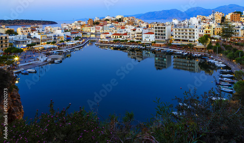 Beautiful southern town of Agios Nikolaos at summer evening. Boats swing on the water of the lake Voulismeni at the pier with evening lights. Crete Island. Greece