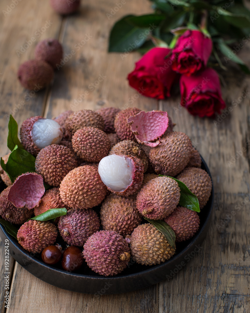 Still life of fresh lychee fruits in a black plate on a wooden background. Rustic style. Decorated with red roses Still life of fresh lychee fruits in a black plate on a wooden background. Rustic style. Decorated with red roses