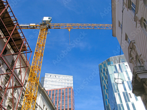 Construction crane on the street. One of the old houses in construction scaffolding and under rebuilding. Arrow of crane in the sky between old buildings. On rear background are modern skyscrapers.