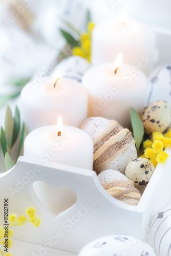 Beautiful Easter composition with white lit candles in a white wooden box with decorated Easter eggs, olive branches and yellow mimosa flowers as a symbol of spring