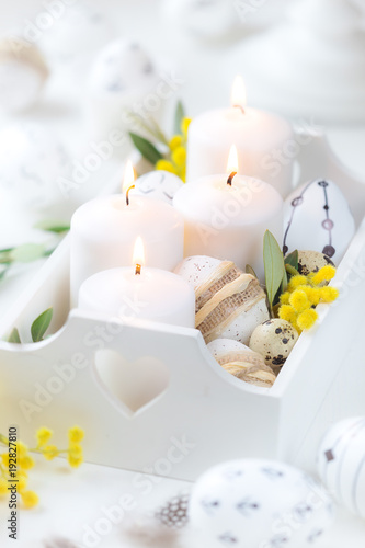 Beautiful Easter composition with white lit candles in a white wooden box with decorated Easter eggs, olive branches and yellow mimosa flowers as a symbol of spring