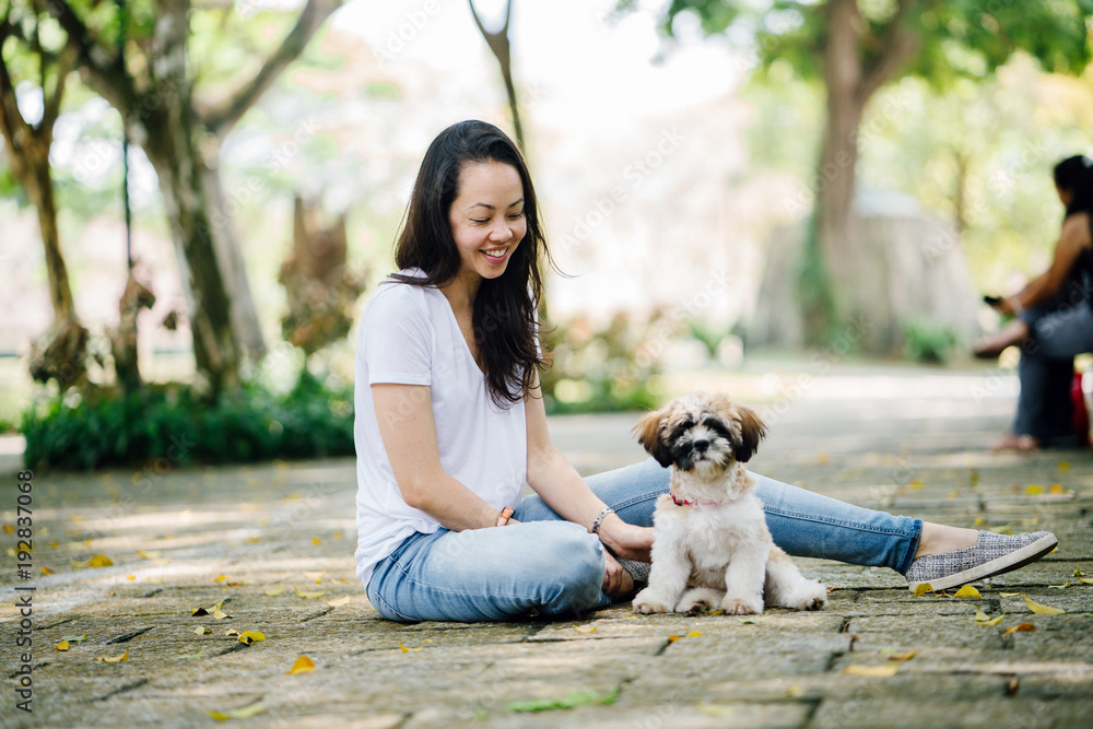 Portrait of young Pan Asian woman with her puppy dog in a park. They are sitting in the park outdoors in the sun during the day and playing with one another. Portrait of young Pan Asian woman with her puppy dog in a park. They are sitting in the park outdoors in the sun during the day and playing with one another.