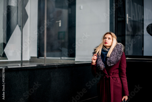 Young girl with blonde hair is standing in a burgundy coat and fur collar near the storefront. Fashion shooting
