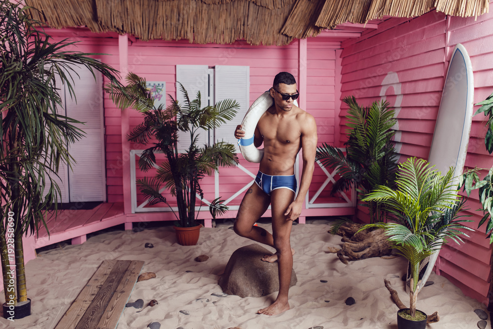 © kanashkin - Sexy african american man in shorts standing on the beach and holding a life buoy.