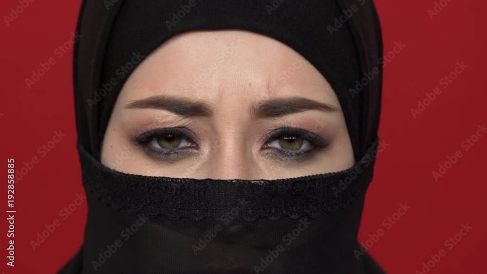 Extreme close up portrait of oriental woman with black smoky eyes wearing traditional niqab expressing anger or frustration, over red background. Concept of emotions Extreme close up portrait of oriental woman with black smoky eyes wearing traditional niqab expressing anger or frustration, over red background. Concept of emotions