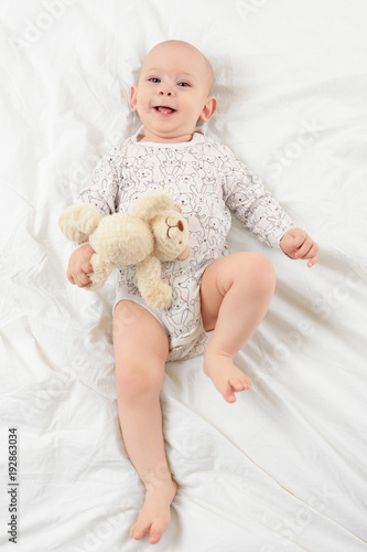 Adorable smiling baby boy with blue eyes lying on a bed with his favorite stuffed teddy bear toy, looking at camera. Cute toddler in pajamas full body portrait.