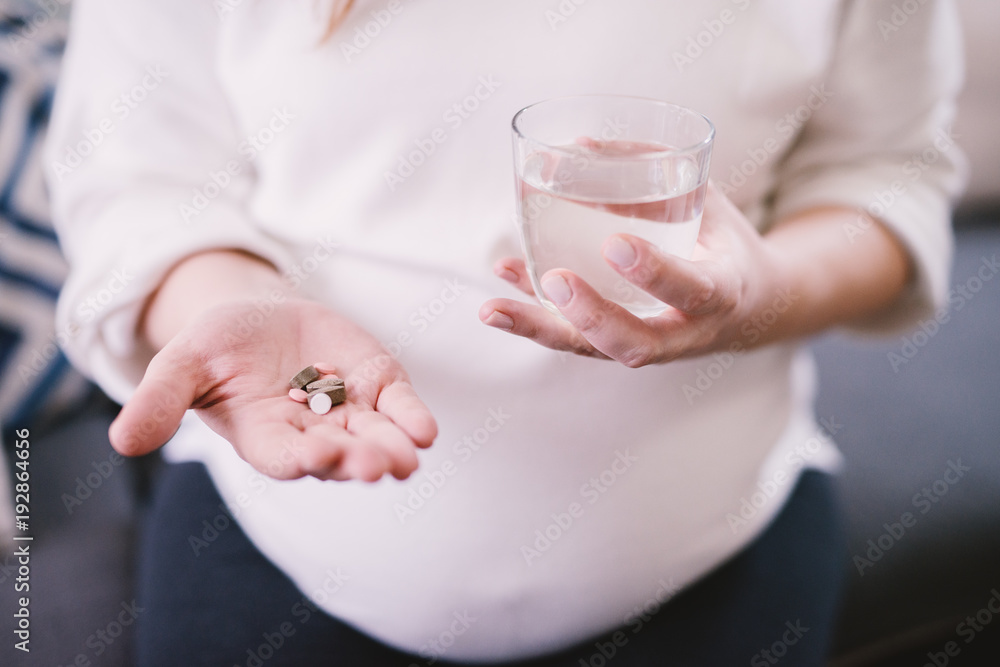 Pregnant girl holding medications and glass of water while sitting on the sofa. Pregnant girl holding medications and glass of water while sitting on the sofa.