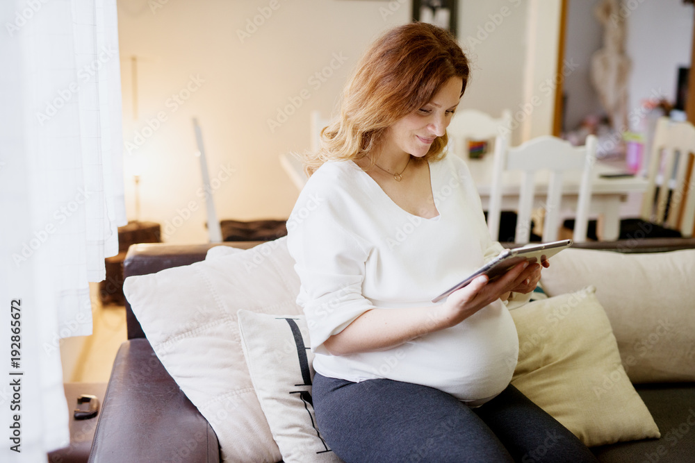 Joyful pregnant woman having fun with a tablet while sitting on sofa at home. Joyful pregnant woman having fun with a tablet while sitting on sofa at home.