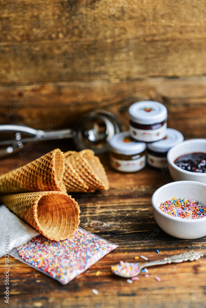 A Kit For Making Ice Cream: Spoon For Ice Cream, Ice Cream Cones, Jam, Topping, Chocolate, Sprinkles, Coconut Shavings. On Wooden Background. The Concept Of A Set, Surprise, Gift Idea A Kit For Making Ice Cream: Spoon For Ice Cream, Ice Cream Cones, Jam, Topping, Chocolate, Sprinkles, Coconut Shavings. On Wooden Background. The Concept Of A Set, Surprise, Gift Idea