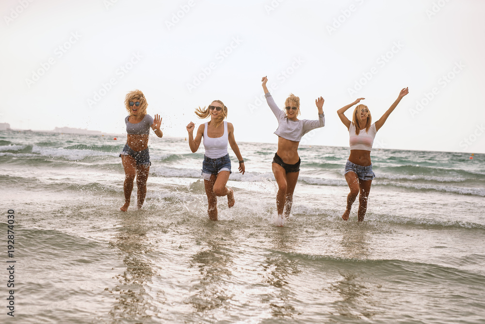 Four sexy ladies running in the sea water and having fun on the beach. - obrazy, fototapety, plakaty 