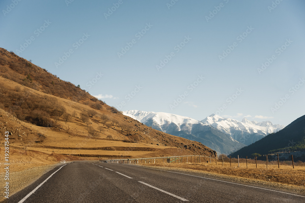Fototapeta premium View of the autumn road leading to the mountains to the snow-capped peaks of the Caucasus. The concept of traveling to the mountains by car